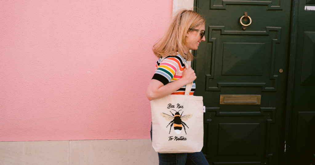 Jane Mosbacher Morris (founder & CEO) walking holding a tote bag featuring a bee reading "Bee Nice to Nature" against a pink wall and green door