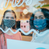 two girls wearing custom cloth face masks behind a glass storefront
